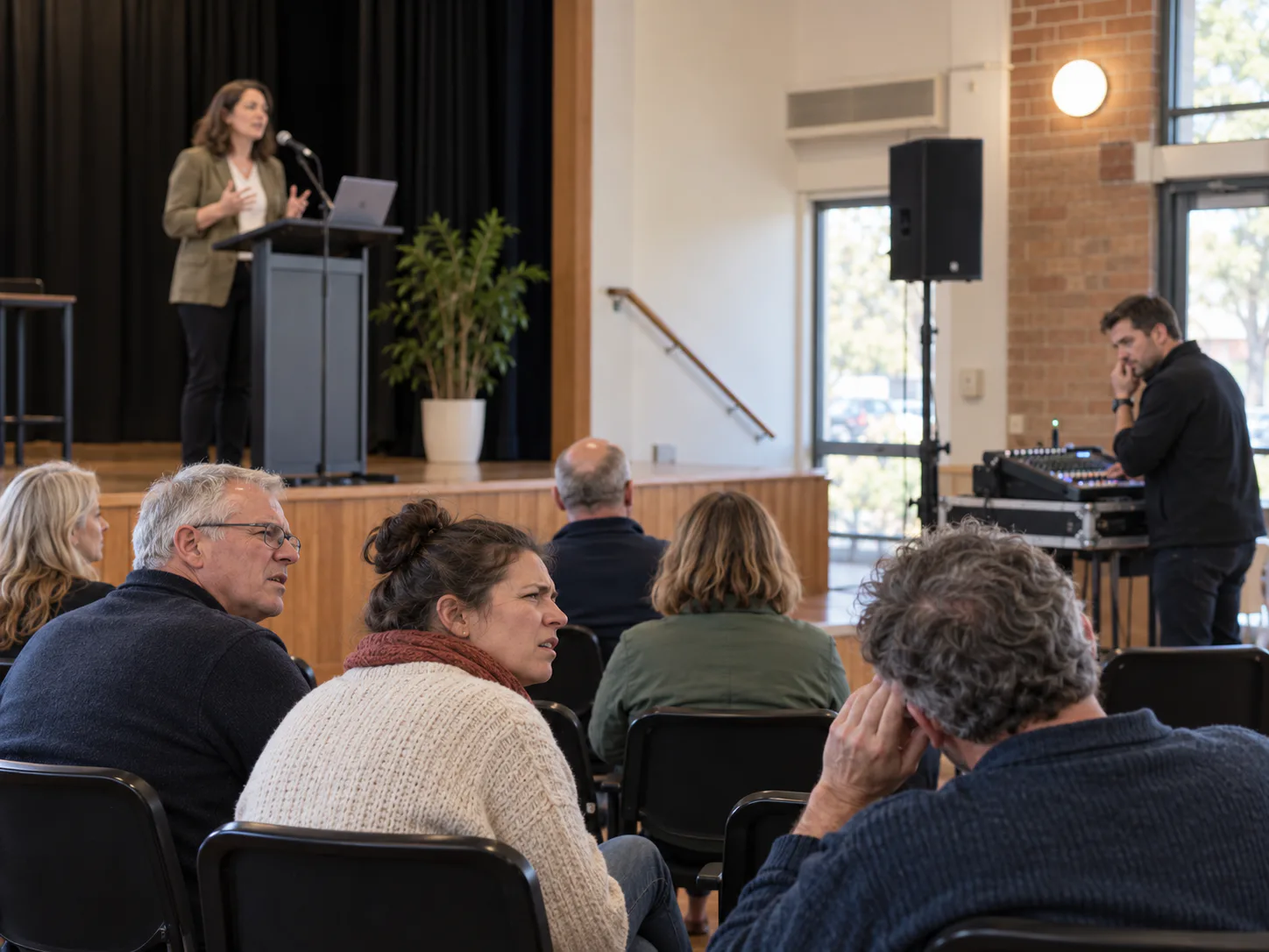 Audience in a hall straining to follow a presenter, one listener with a hand to her ear, speakers mounted high on the wall