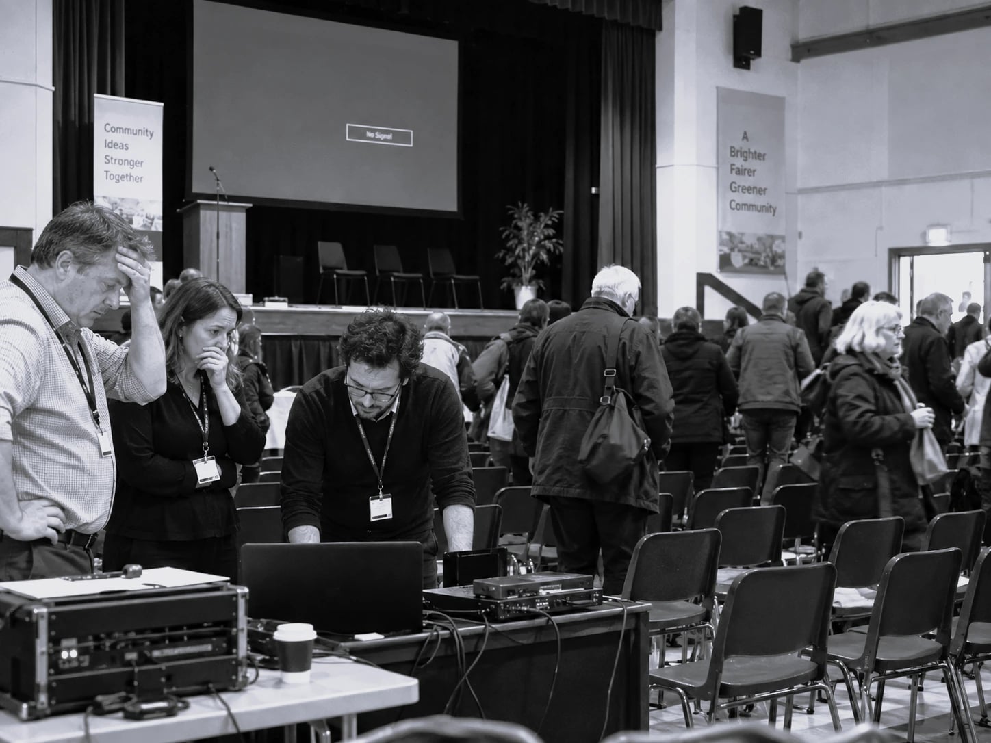 Staff troubleshooting AV equipment at a desk in a hall as the audience leaves, screen showing No Signal