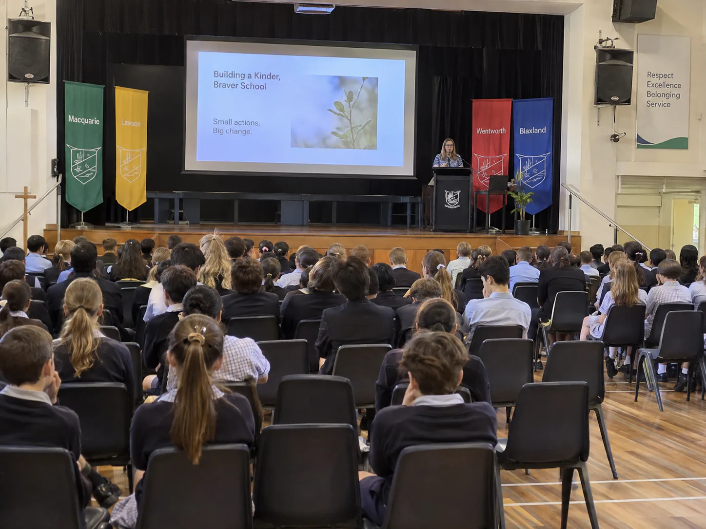Students seated in a school hall during an assembly, presenter at a lectern with projection screen and wall speakers