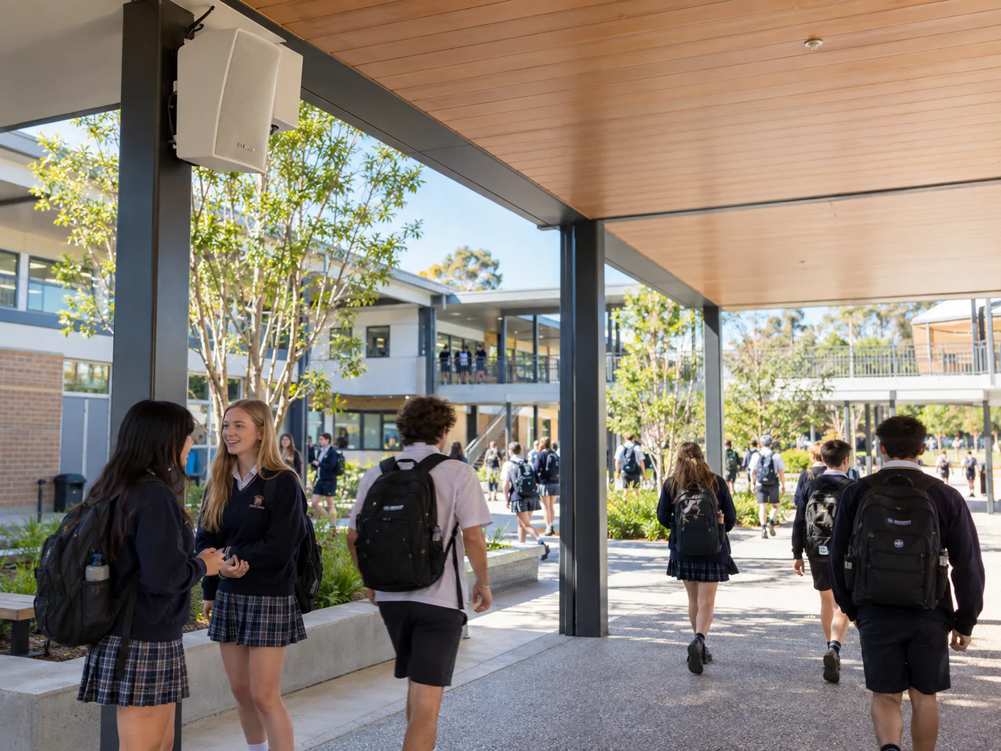 Students moving through a covered school walkway beneath a wall-mounted PA speaker